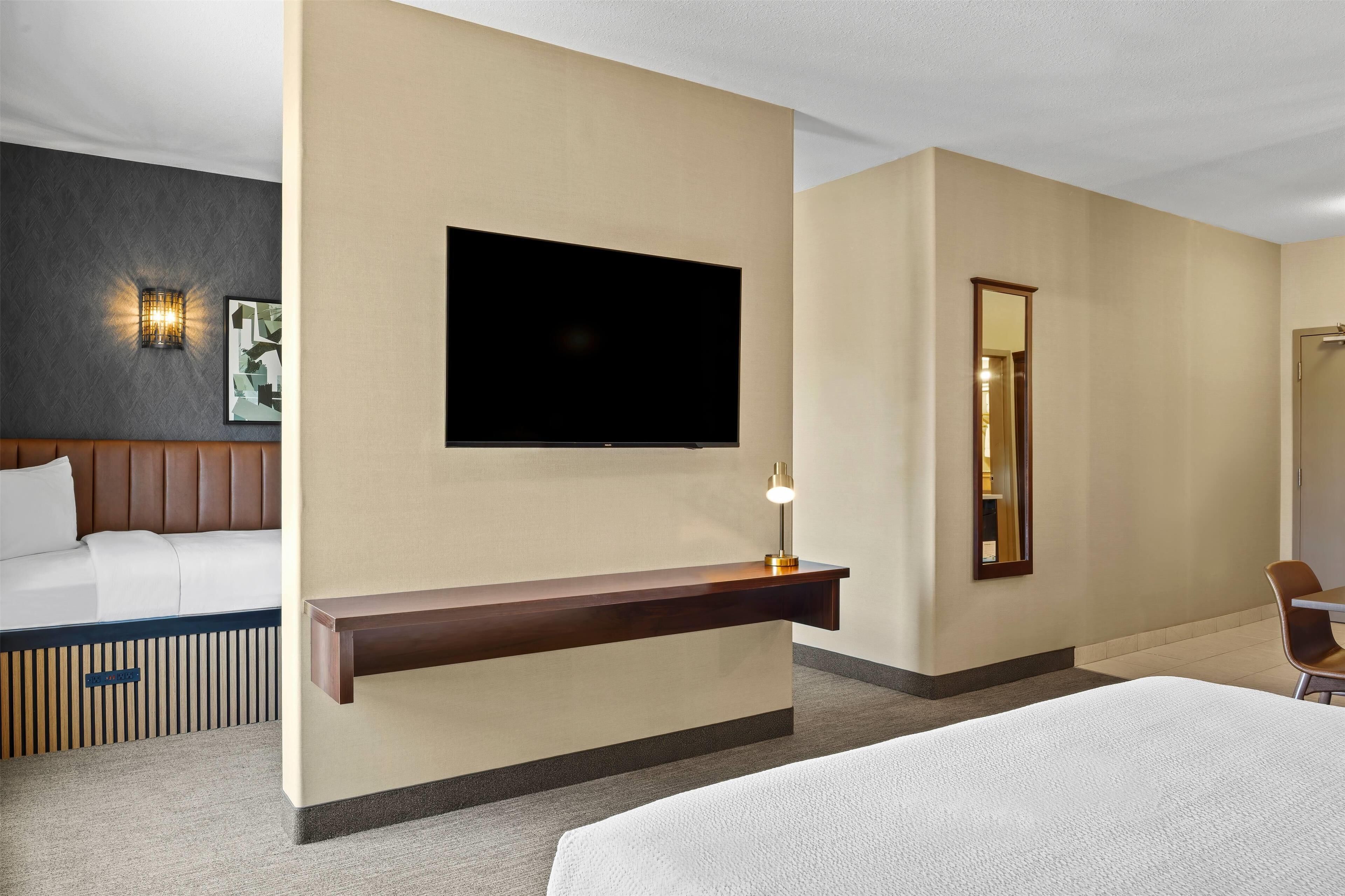 A hotel room with a bed, a flat-screen TV mounted on the wall, a bedside table with a lamp, a mirror, and a seating area with a chair.