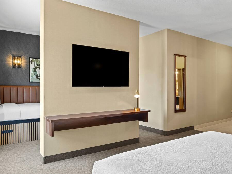 Quarto de hotel com cama, televisão de tela plana na parede, mesa de cabeceira com abajur, espelho e área de estar com uma poltrona.