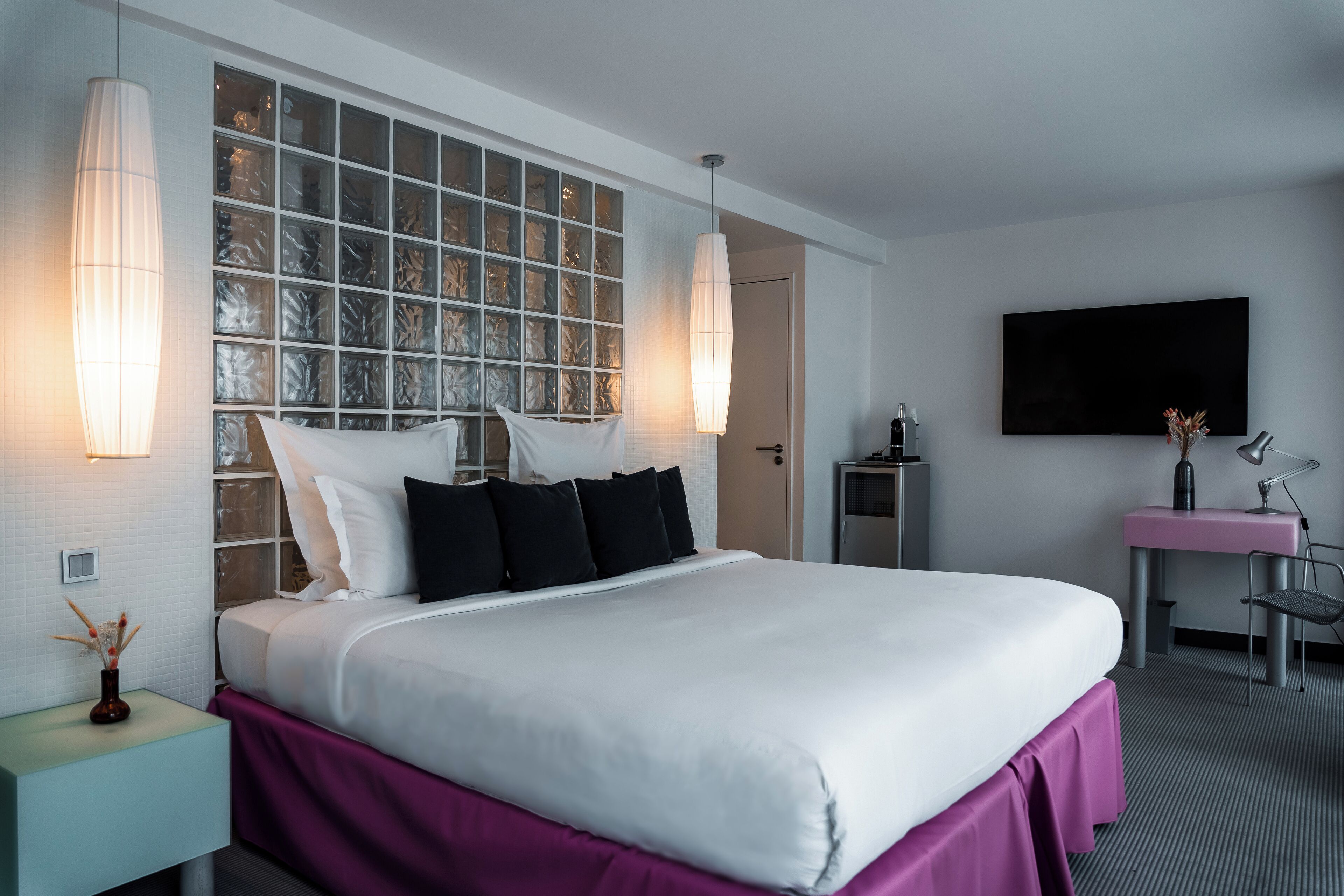 A modern hotel room with a large bed, a desk with a chair, a flat-screen TV, and a decorative wall with glass panels.