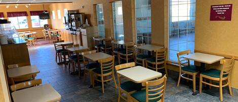 A restaurant with wooden tables and chairs, a kitchen area, and large windows.
