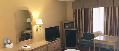 A hotel room with a bed, a desk with a lamp, a television, and a chair.