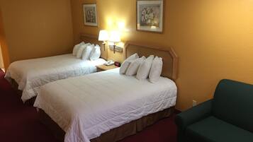 A hotel room with two beds, a nightstand with a lamp, a green armchair, and two framed floral paintings on the wall.