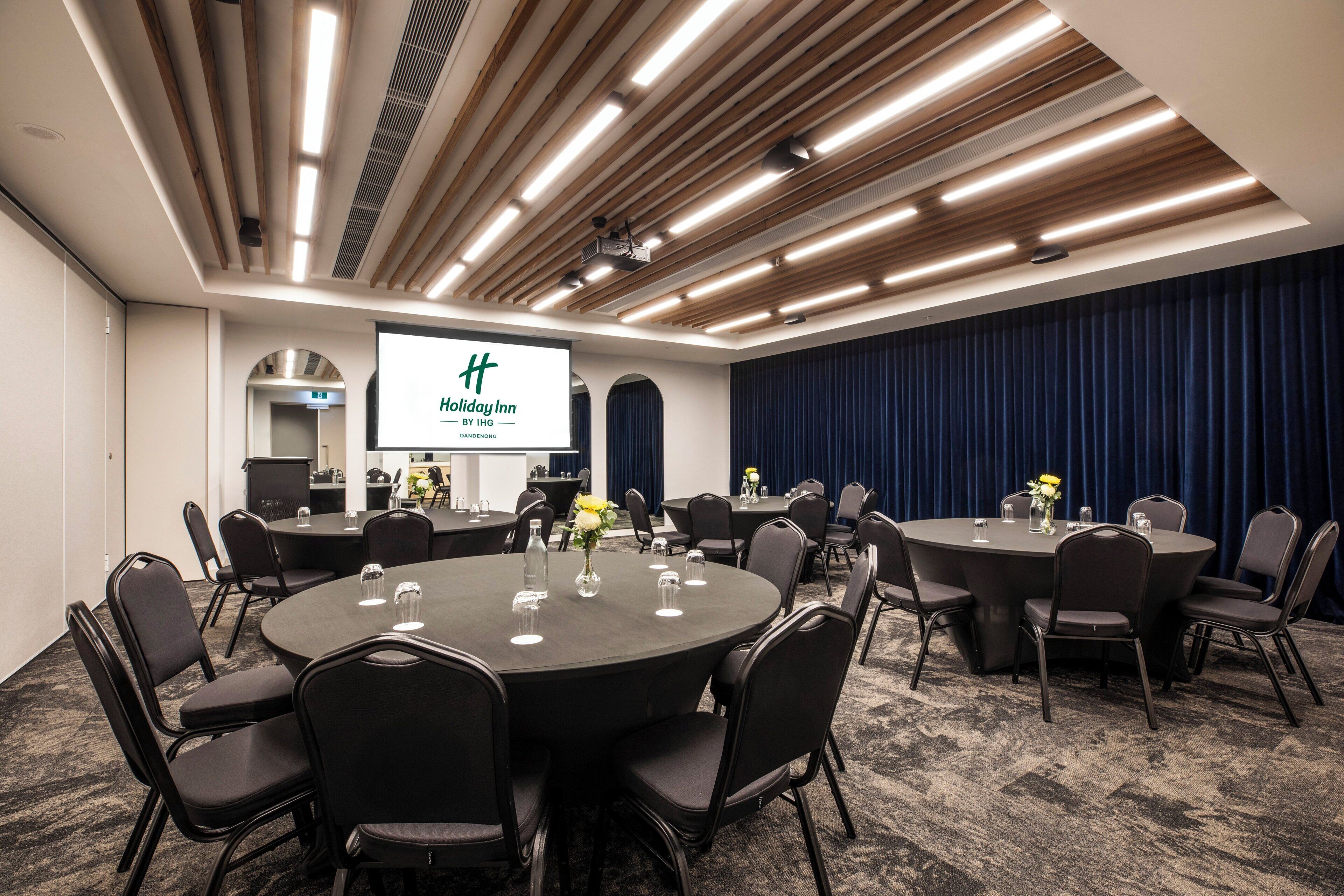 A conference room with multiple tables and chairs, a projector screen displaying 'Holiday Inn by IHG', and a dark blue curtain backdrop.
