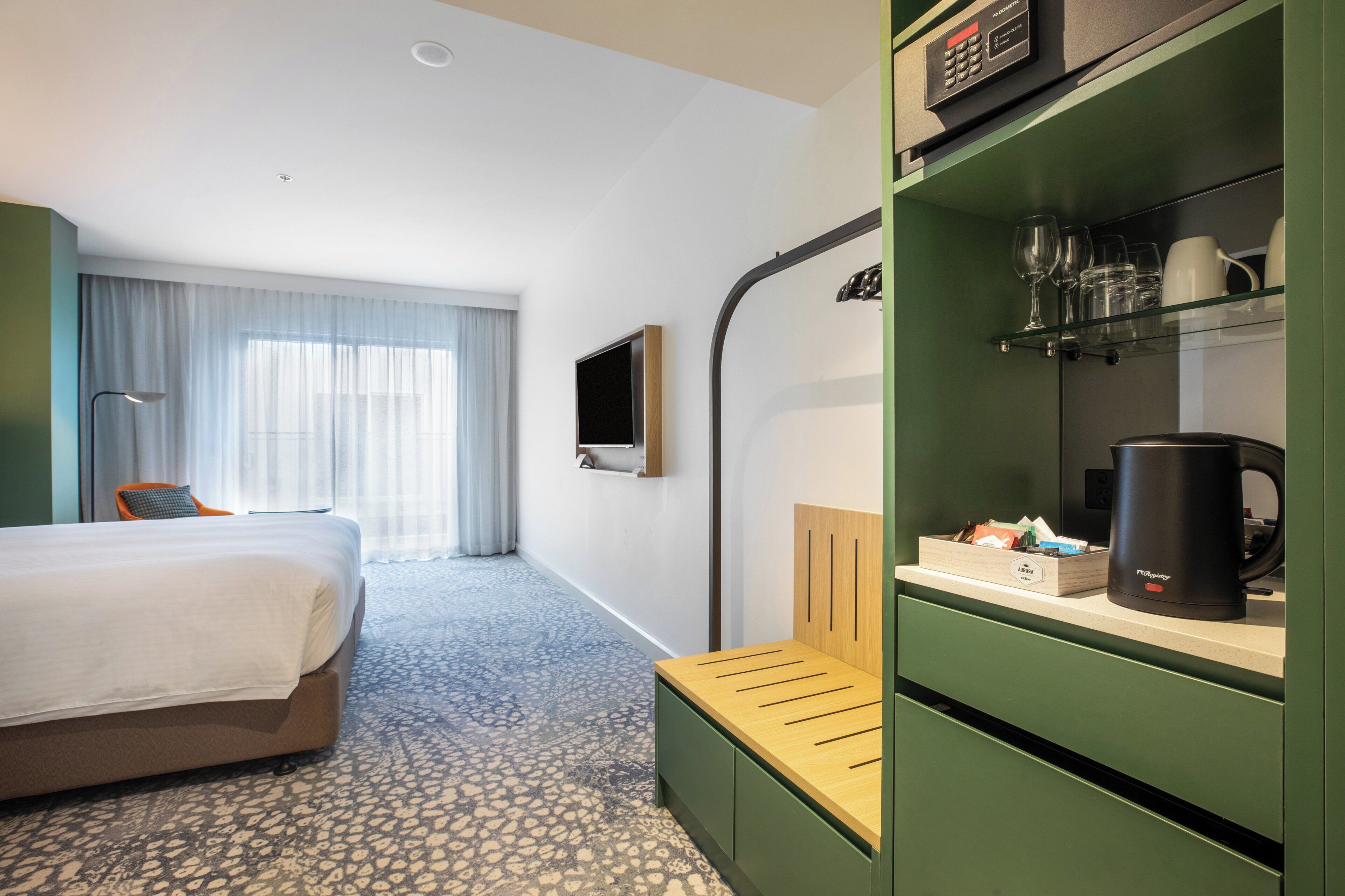 Una habitación de hotel con cama, escritorio con cafetera, televisor y estante con vasos y tazas.