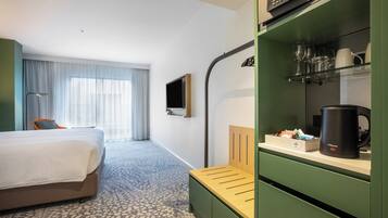 Una habitación de hotel con cama, escritorio con cafetera, televisor y estante con vasos y tazas.