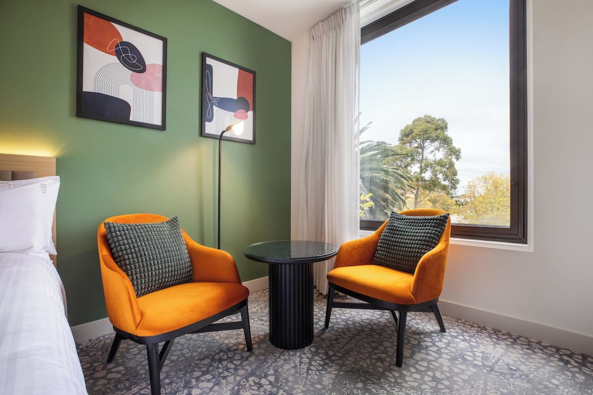 Une chambre d’hôtel avec deux fauteuils orange, une petite table ronde et un lit avec une literie blanche. Deux œuvres d’art encadrées ornent le mur, et une grande fenêtre offre une vue sur les arbres.