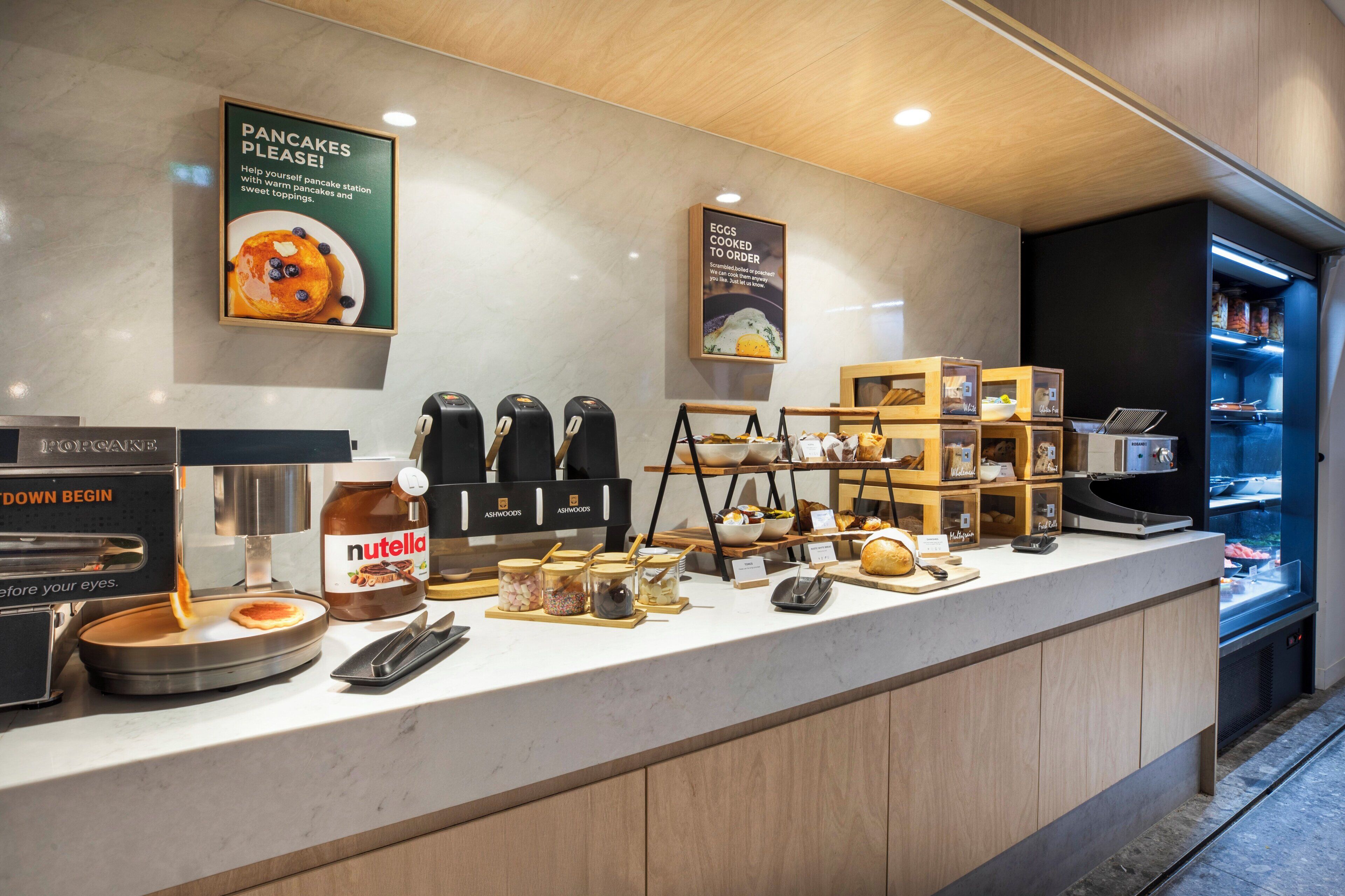 Estação de pequeno-almoço com dispensador de Nutella, máquinas de café e placa a indicar panquecas.