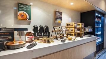 Estação de pequeno-almoço com dispensador de Nutella, máquinas de café e placa a indicar panquecas.