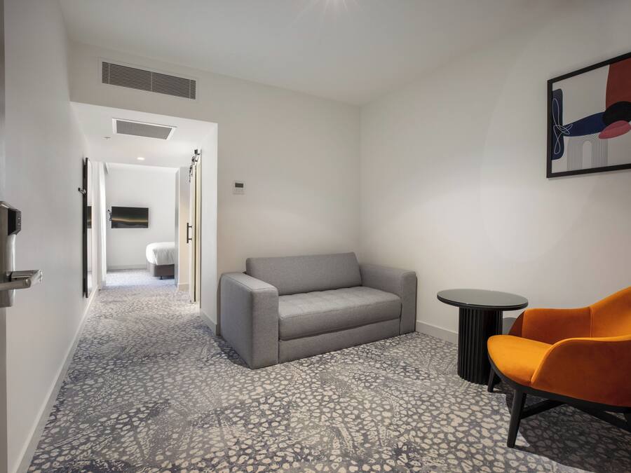 A hotel room with a grey sofa, an orange armchair, a small round table, and a patterned floor.