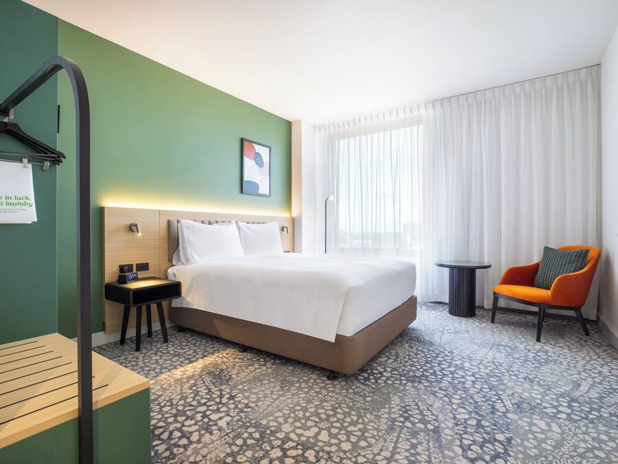 Una habitación de hotel con cama, una silla, una mesita y un perchero en la pared.