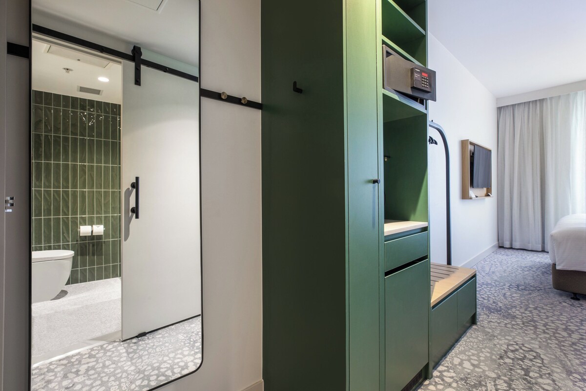 Una habitación de hotel con un armario verde, un baño con inodoro y ducha, y una cama con cabecero.