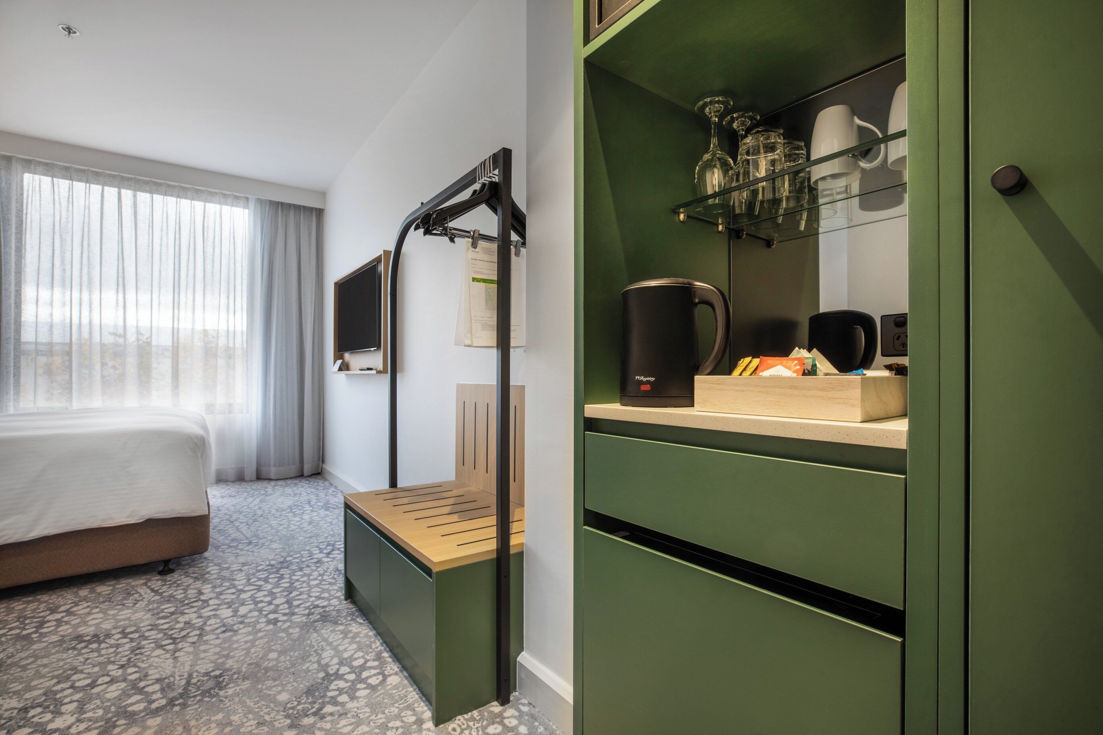 A hotel room with a bed, a bench, a desk with a coffee maker, and a green cabinet with shelves.