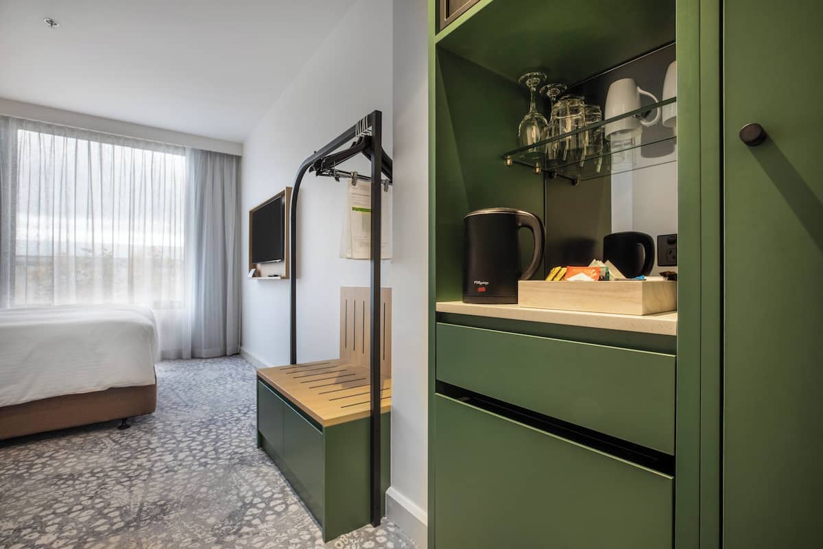 Habitación de hotel con cama, un banco, un escritorio con cafetera y un mueble verde con estantes.