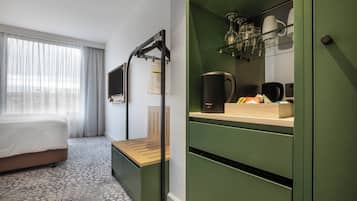 A hotel room with a bed, a bench, a desk with a coffee maker, and a green cabinet with shelves.
