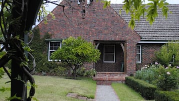 Uma casa de tijolos com varanda frontal, uma pequena árvore e um gramado bem cuidado.