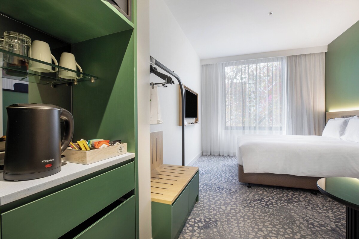 Una habitación de hotel con una cama, una cafetera, un escritorio con portavasos y una ventana con cortinas.