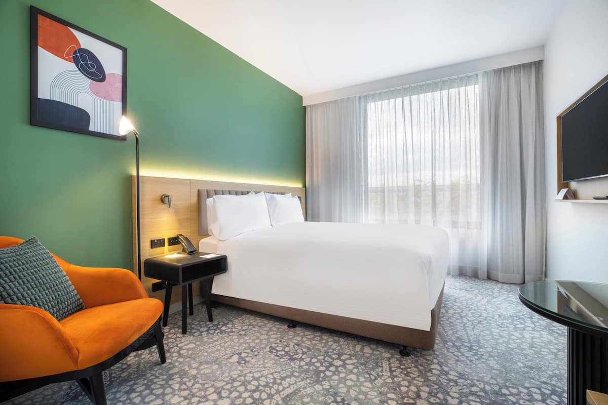 Une chambre d’hôtel avec un lit, une table de chevet, une lampe, une chaise et une grande fenêtre avec des rideaux.