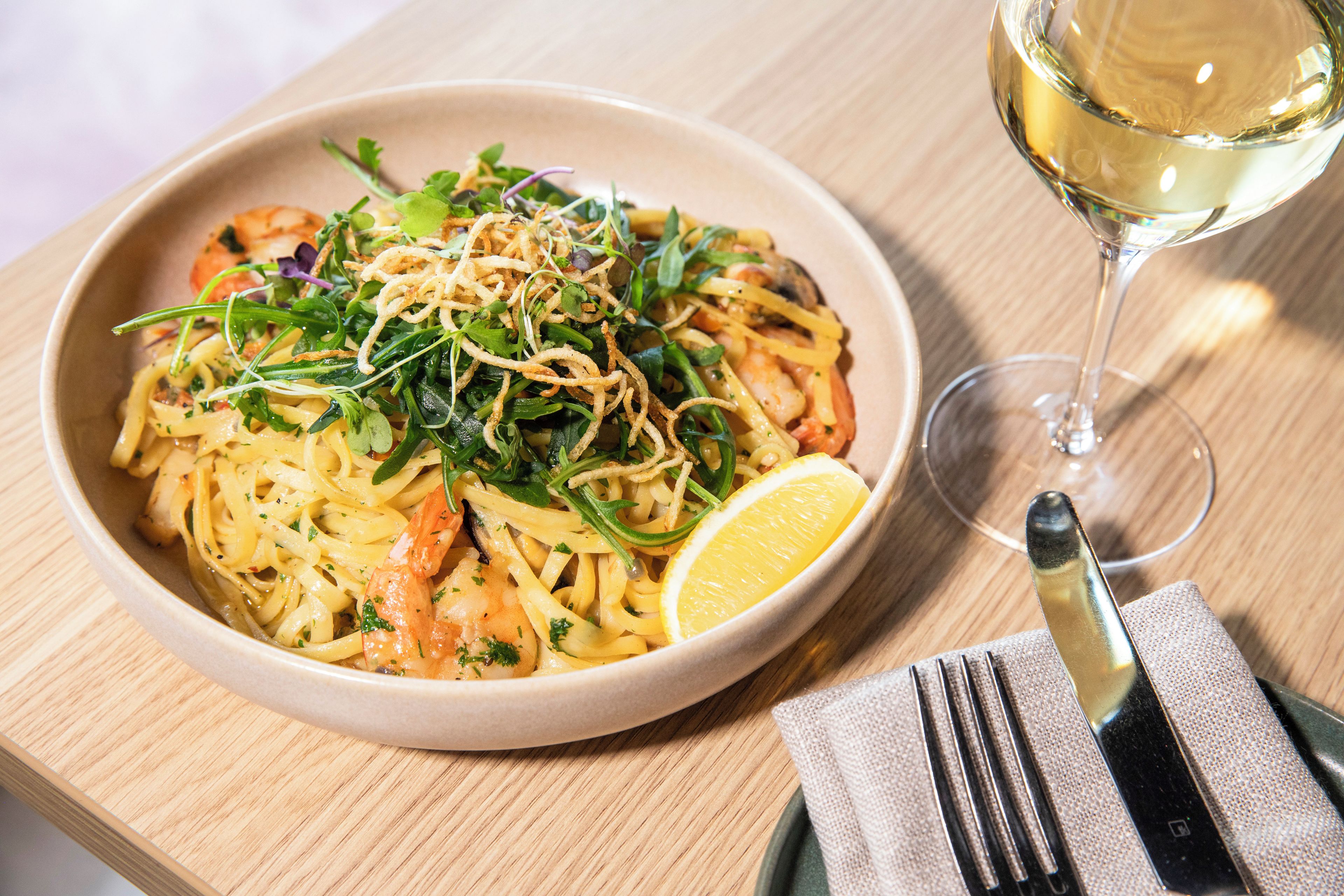 Un plato de pasta con camarones, adornado con vegetales verdes y limón, sobre una mesa de madera con una copa de vino blanco y cubiertos.
