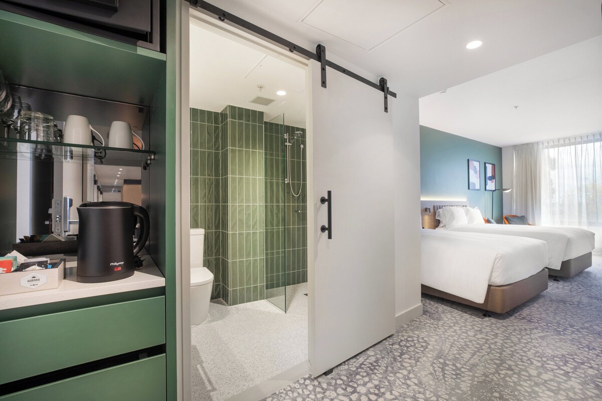 Habitación de hotel con puerta corredera que da a zona de ducha, cama y cafetera sobre la encimera.