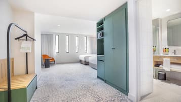 Ein Hotelzimmer mit einem Bett, einer Sitzecke mit einem Sessel, einem Kleiderschrank, einem Badezimmer mit Spiegel und Pflegeartikeln sowie einem Fenster mit Vorhängen.