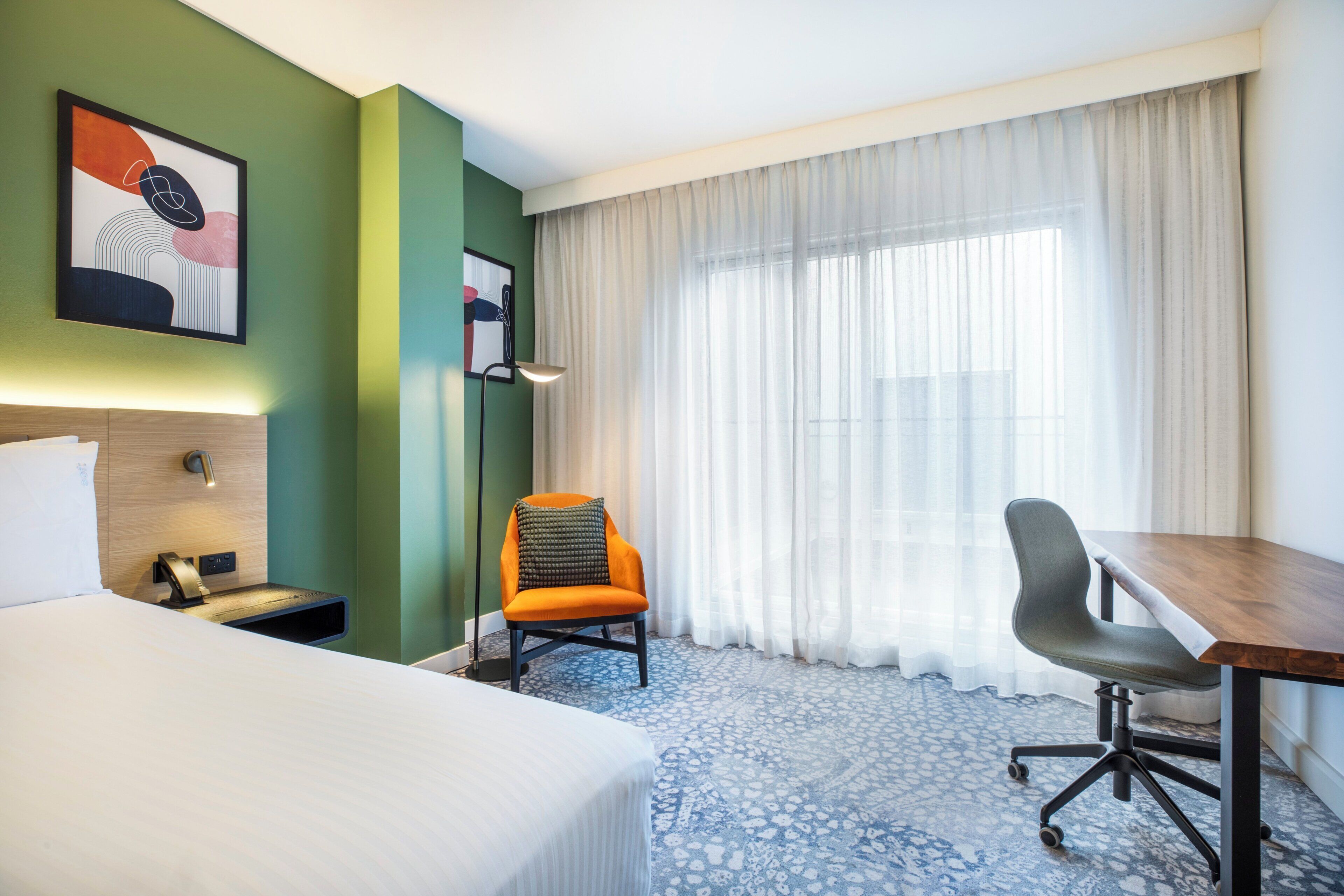 A hotel room with a bed, a desk, an orange chair, a lamp, and a large window with sheer curtains.