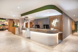 A modern reception desk with a curved design, a wooden panel wall, and a sign with a light-up 'H' logo. - Holiday Inn Dandenong by IHG (Dandenong)