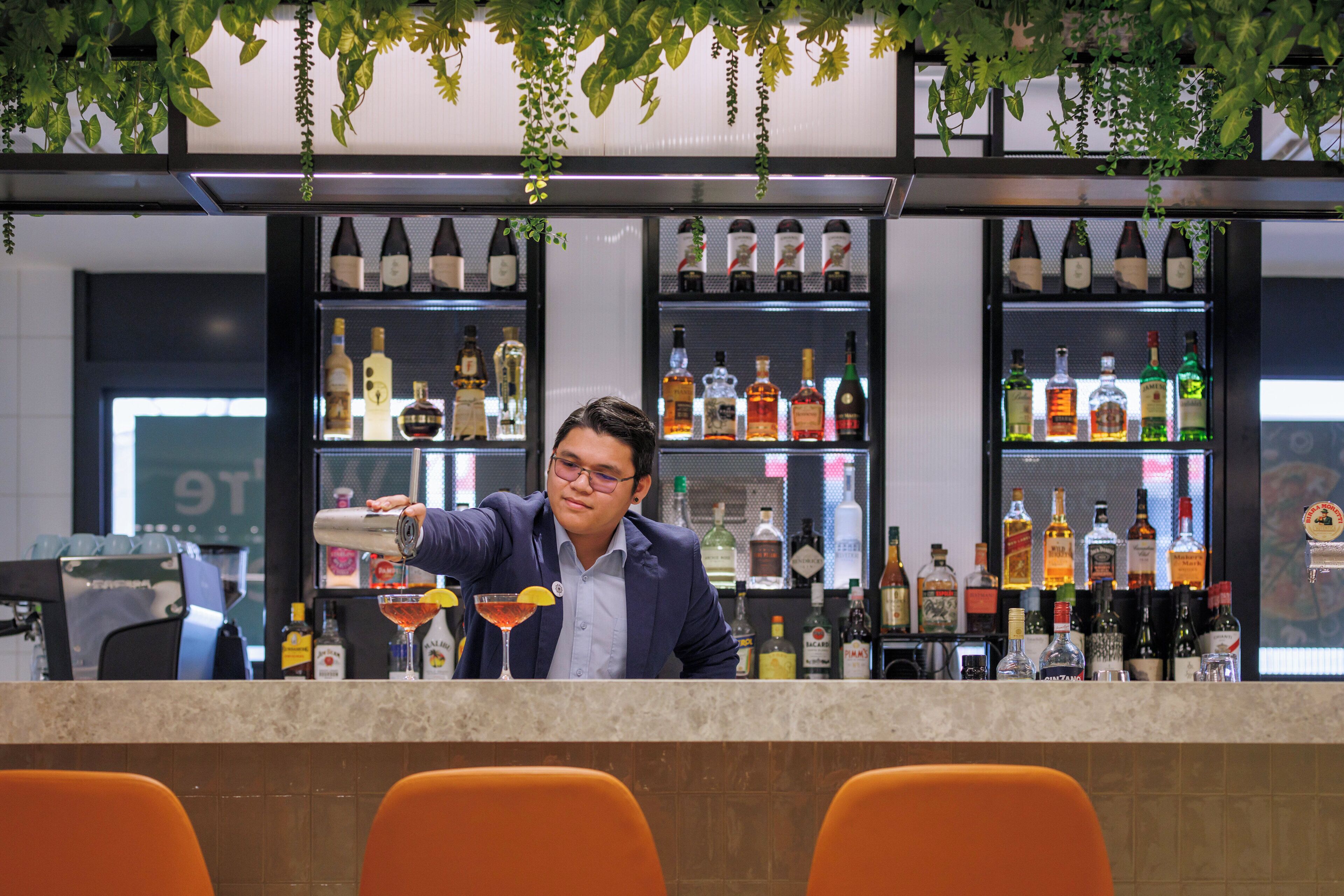 A bartender preparing a drink behind a bar with shelves of various liquor bottles.