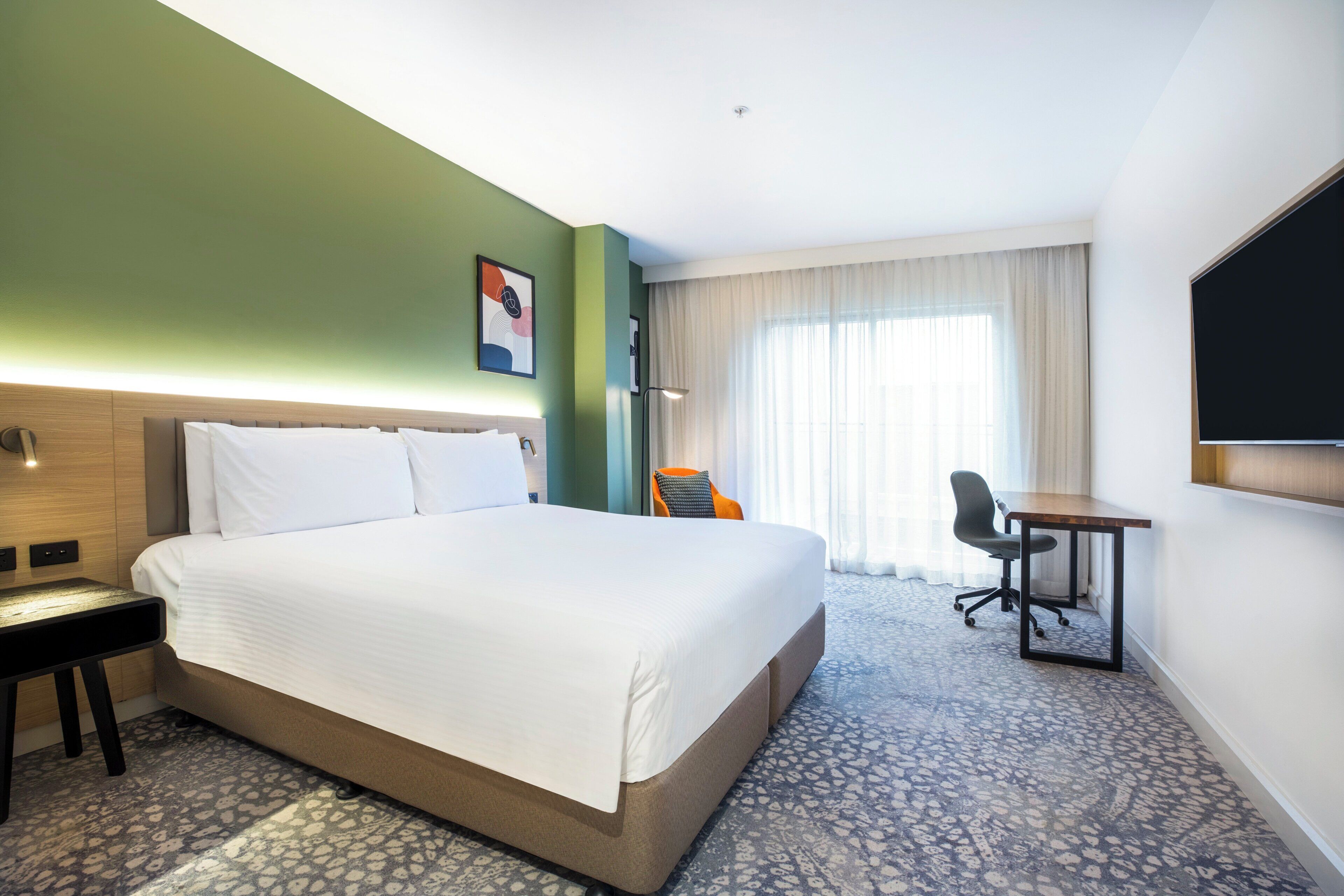 Una habitación de hotel con una cama grande, un escritorio, una silla, un televisor de pantalla plana y un ventanal con cortinas.