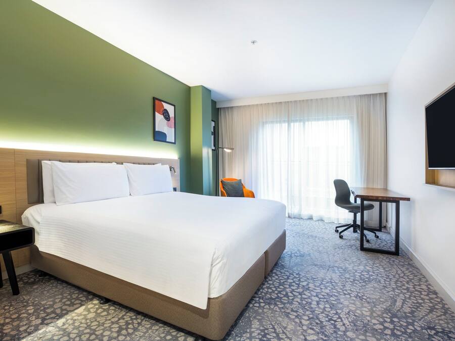 Una habitación de hotel con una cama grande, un escritorio, una silla, un televisor de pantalla plana y un ventanal con cortinas.