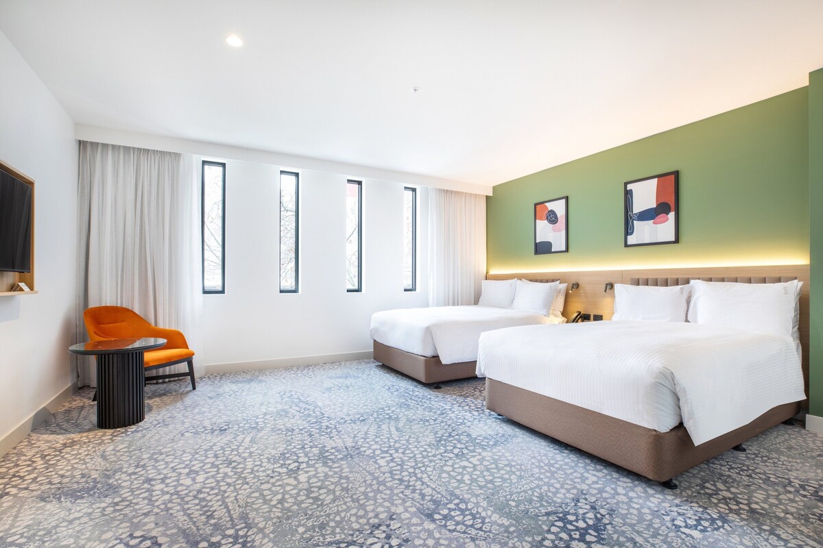 Habitación de hotel con dos camas, una silla, una mesita pequeña y un ventanal con cortinas.
