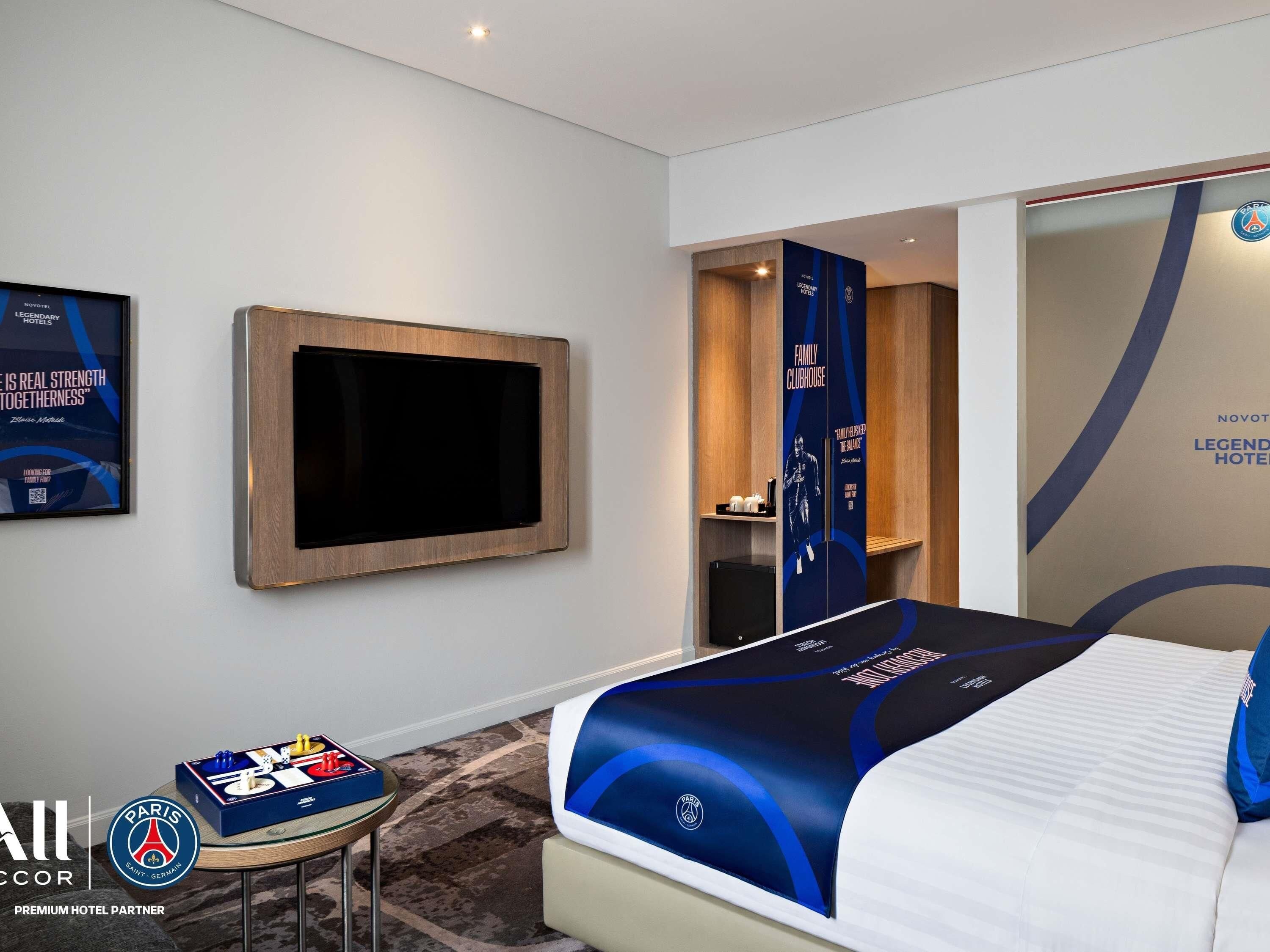 A hotel room with a large bed, a flat-screen TV mounted on the wall, a minibar, and a view of the hallway.