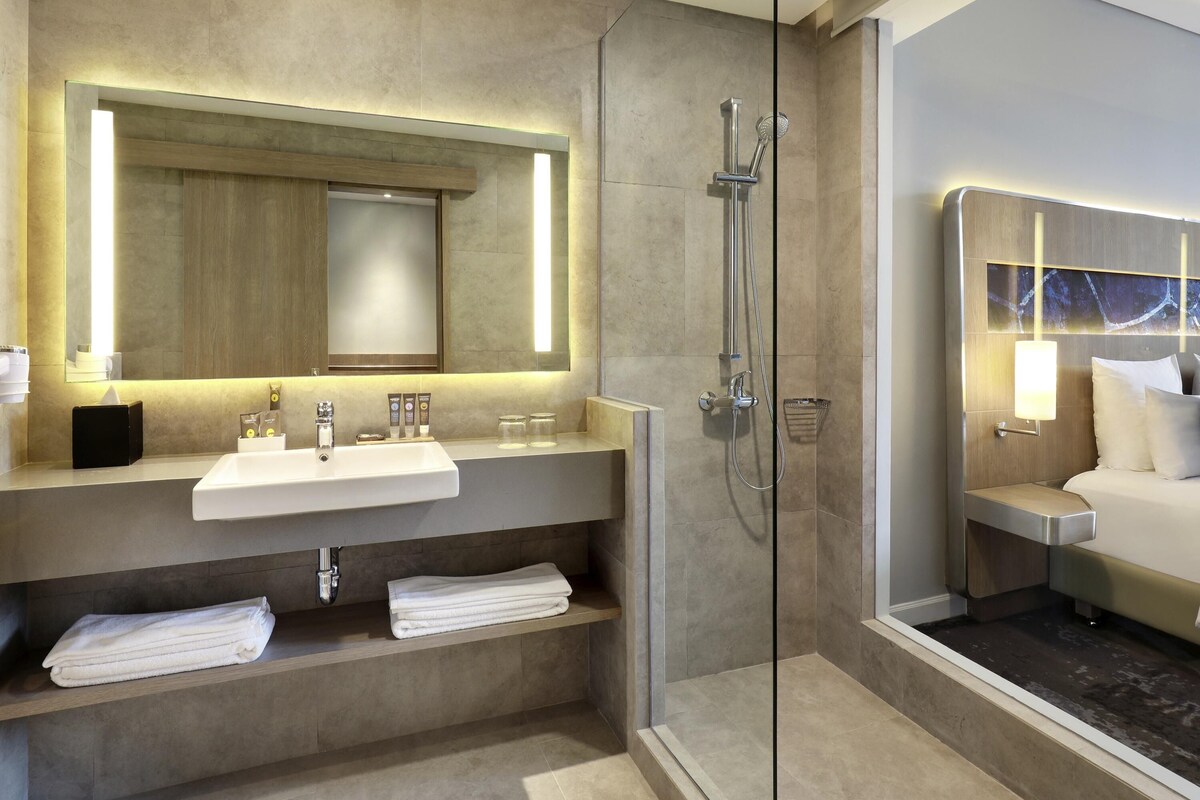 Une salle de bain moderne avec une douche à parois de verre, un meuble-lavabo avec un évier et un miroir, et un espace chambre séparé comprenant un lit et une table de chevet.