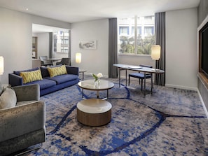 Ein modernes Hotelzimmer mit blauem Teppich, einem runden Couchtisch, einer Couch, einem Sessel, einem Esstisch und einem großen Fenster mit Blick auf Gebäude und Bäume.