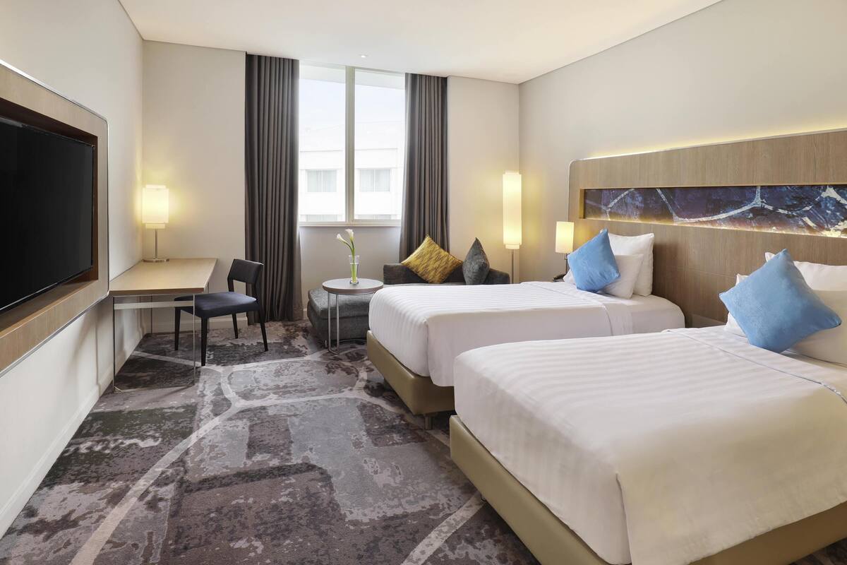A hotel room with two beds, a desk, a chair, a TV, and a window with curtains.