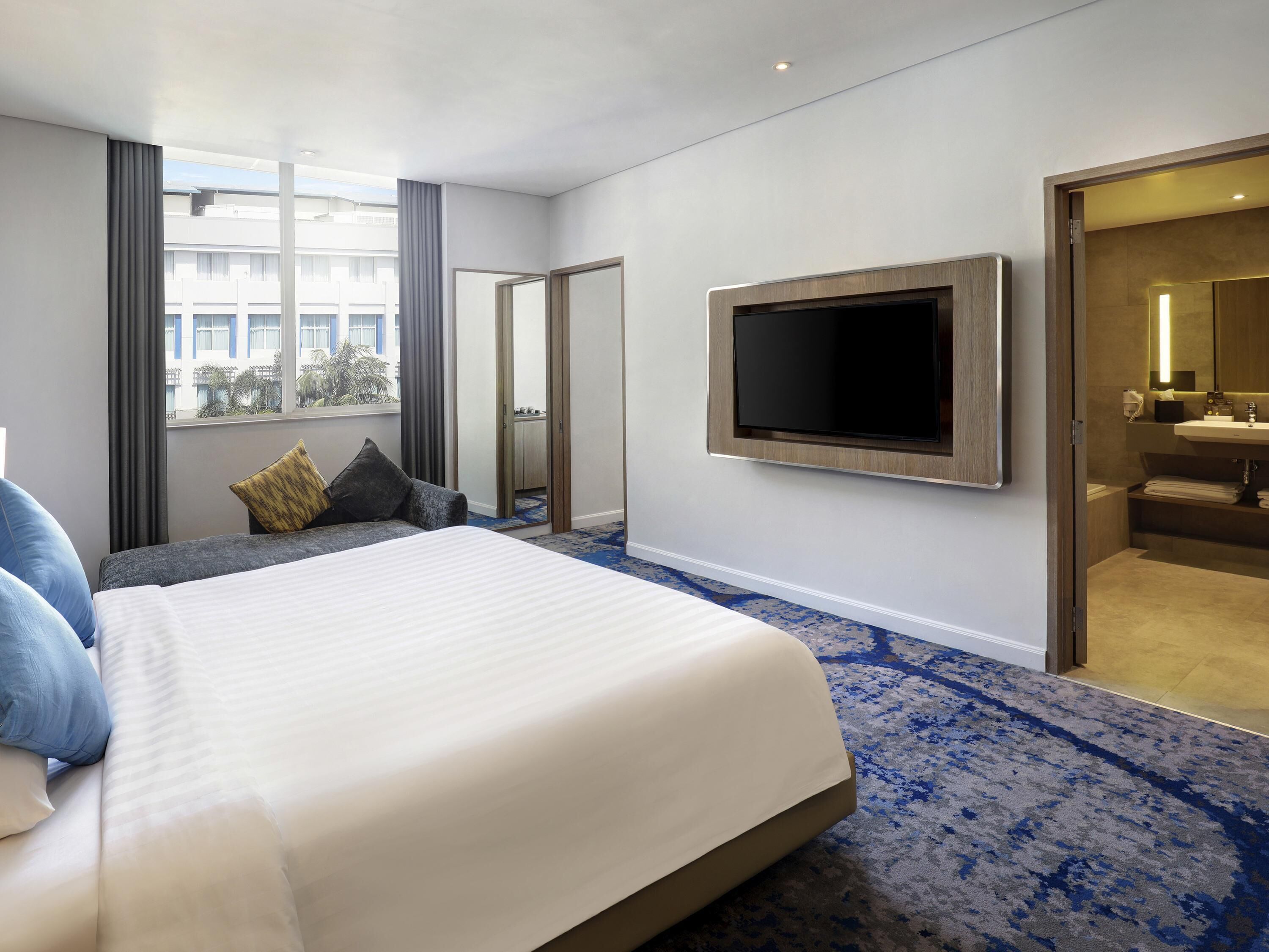 Habitación de hotel con una cama grande, un televisor de pantalla plana montado en la pared, un baño con lavamanos y espejo, y vistas de la ciudad a través de la ventana.