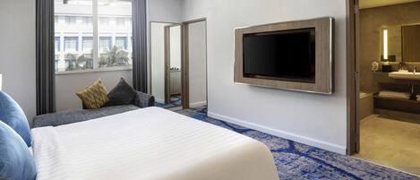 Habitación de hotel con una cama grande, un televisor de pantalla plana montado en la pared, un baño con lavamanos y espejo, y vistas de la ciudad a través de la ventana.