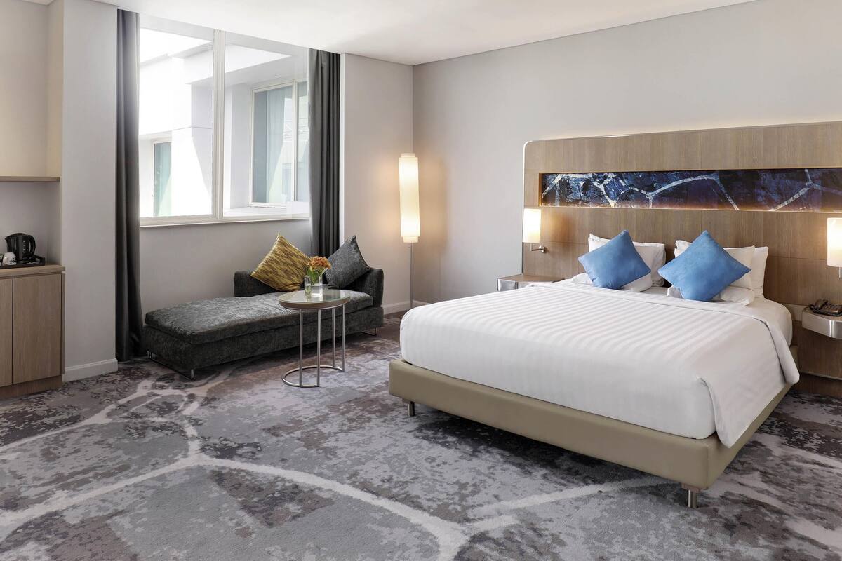 A hotel room with a bed, a sofa, a small table, a lamp, and a view of the outside.