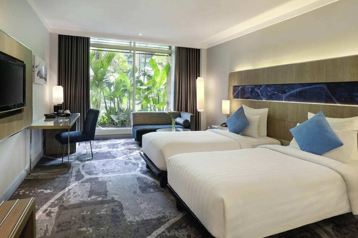 A hotel room with two beds, a desk, a chair, a TV, and large windows with a view of greenery.
