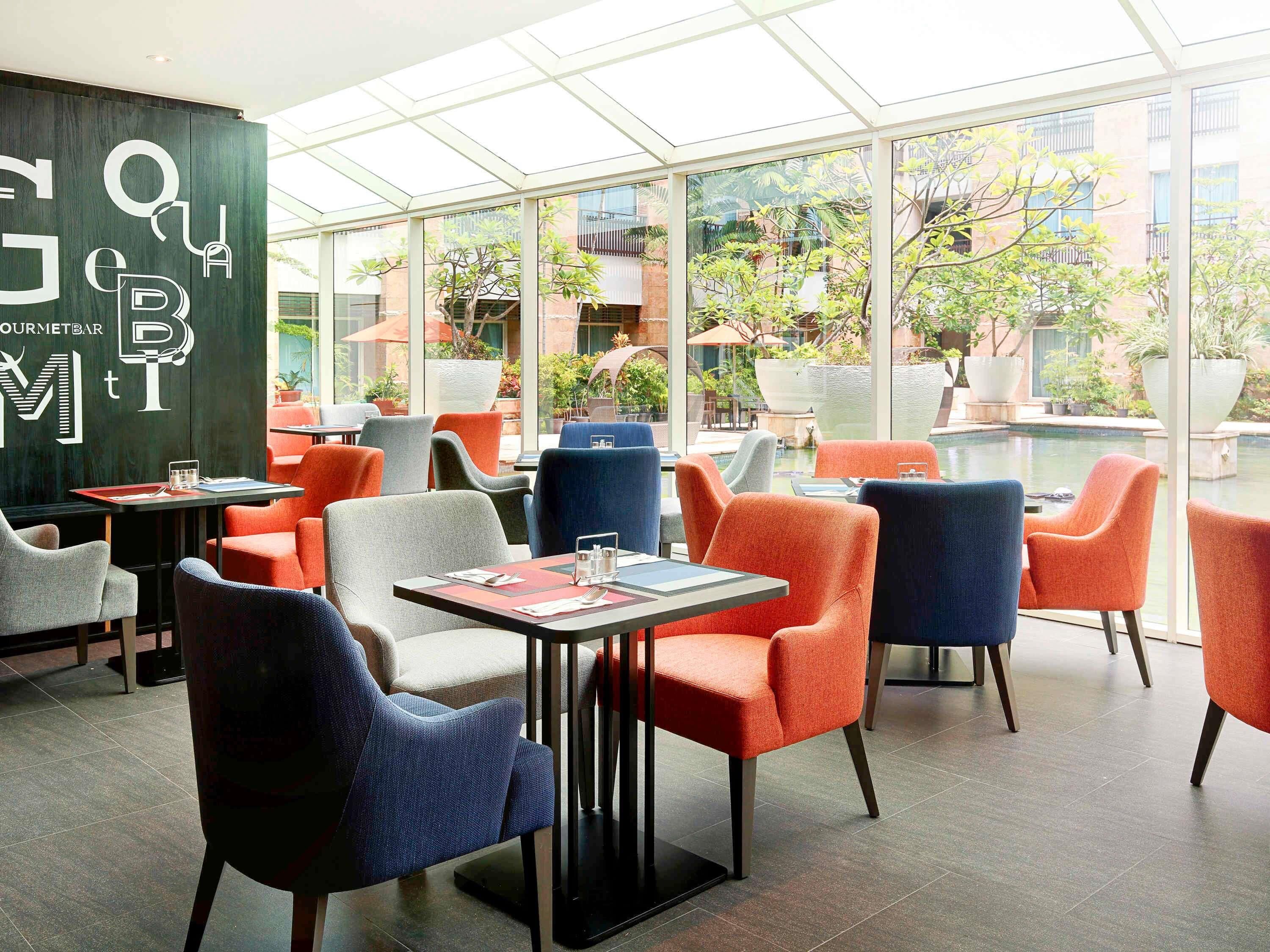A modern outdoor seating area with colorful chairs and tables, large windows offering views of a courtyard with plants and a pool.
