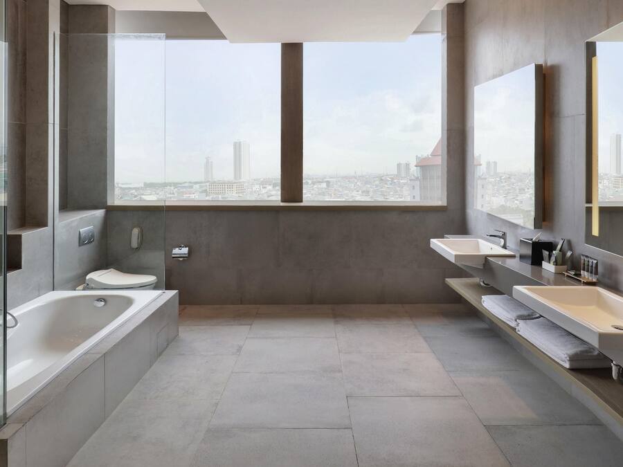 Une salle de bain moderne équipée d’une baignoire, de toilettes et de deux lavabos, avec une grande fenêtre offrant une vue sur la ville.