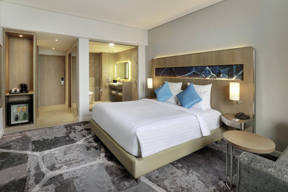 A modern hotel room with a large bed, a bedside table, a chair, a minibar, and a bathroom visible through an open door.