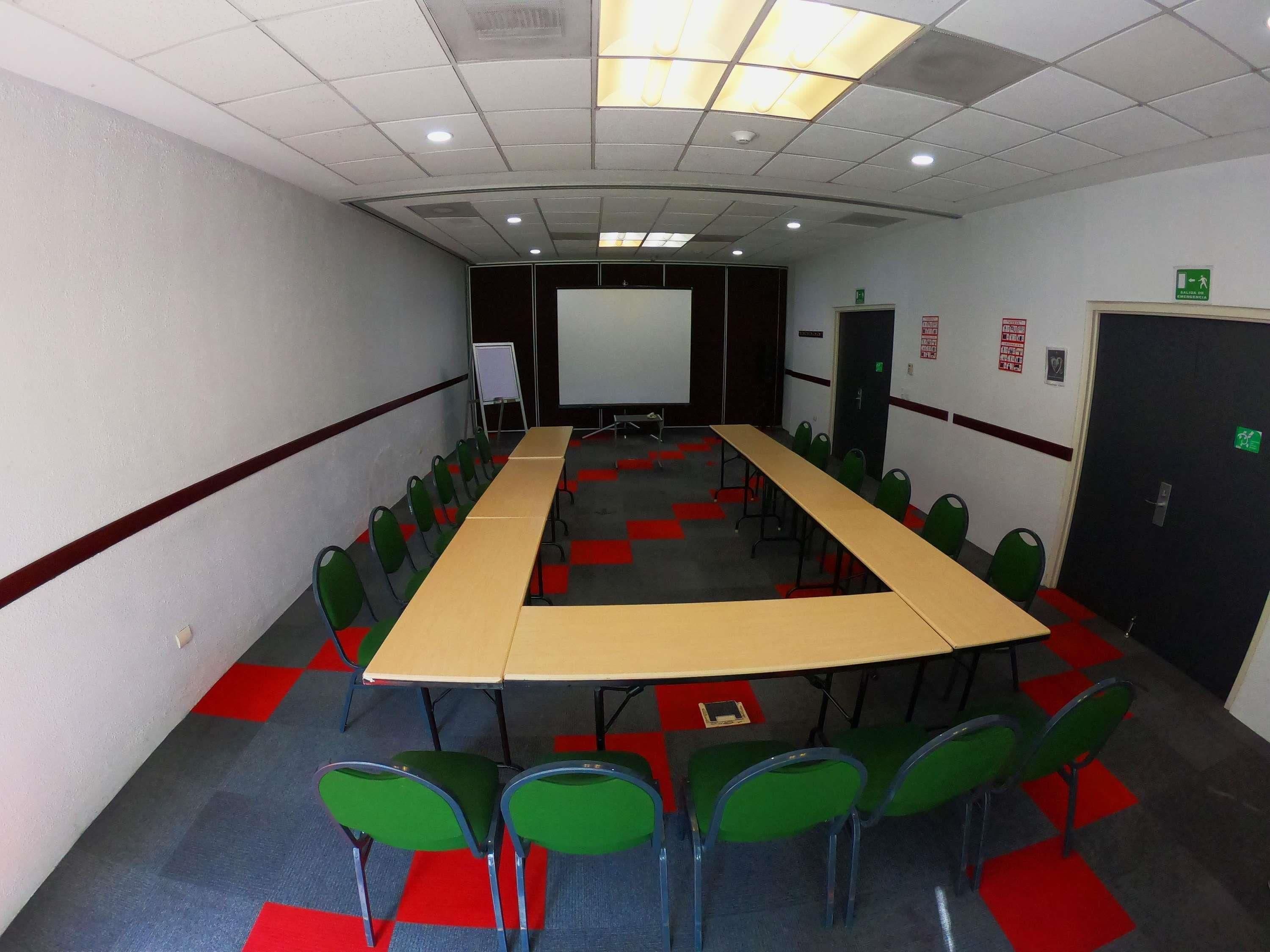 Un salón de conferencias con una larga mesa, sillas verdes, una pizarra y una pantalla de proyector.