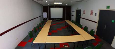 Un salón de conferencias con una larga mesa, sillas verdes, una pizarra y una pantalla de proyector.