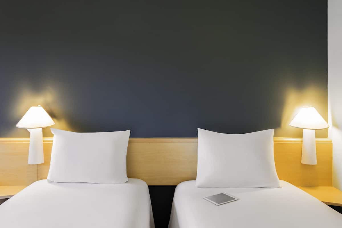 Two single beds with white linens, each with a bedside lamp and a small rectangular object on the bed.