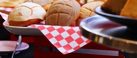 A food display with various baked goods, including bread rolls and a cake, on a table with a checkered paper liner.