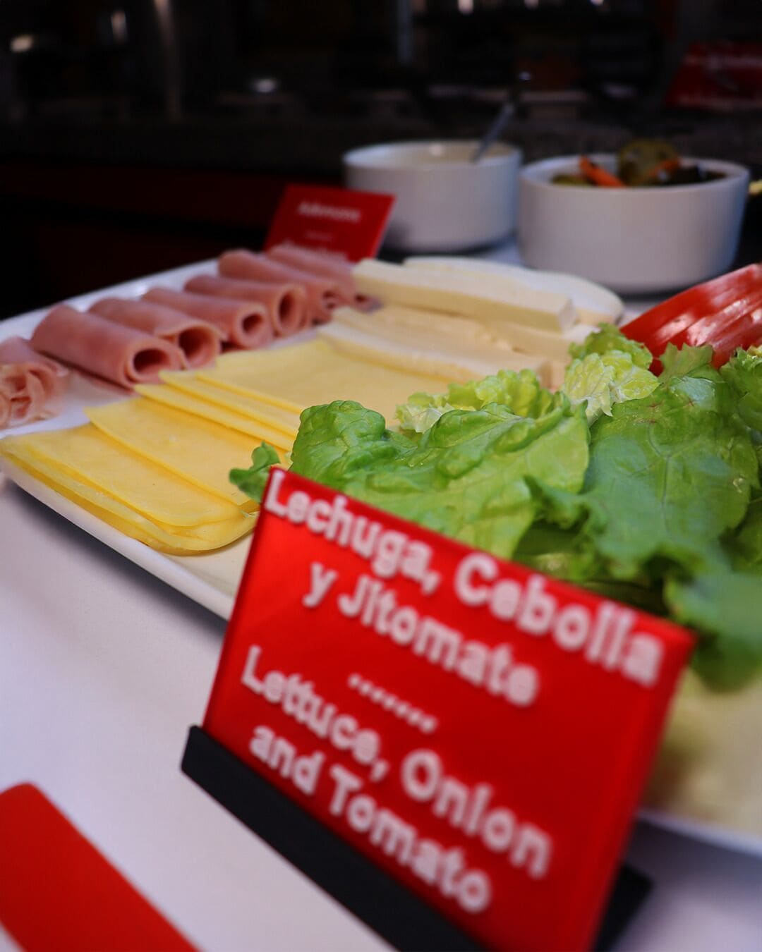 Une table de buffet proposant divers aliments, notamment du jambon, du fromage et de la laitue, ainsi qu’un panneau indiquant la présence de laitue, d’oignon et de tomate.
