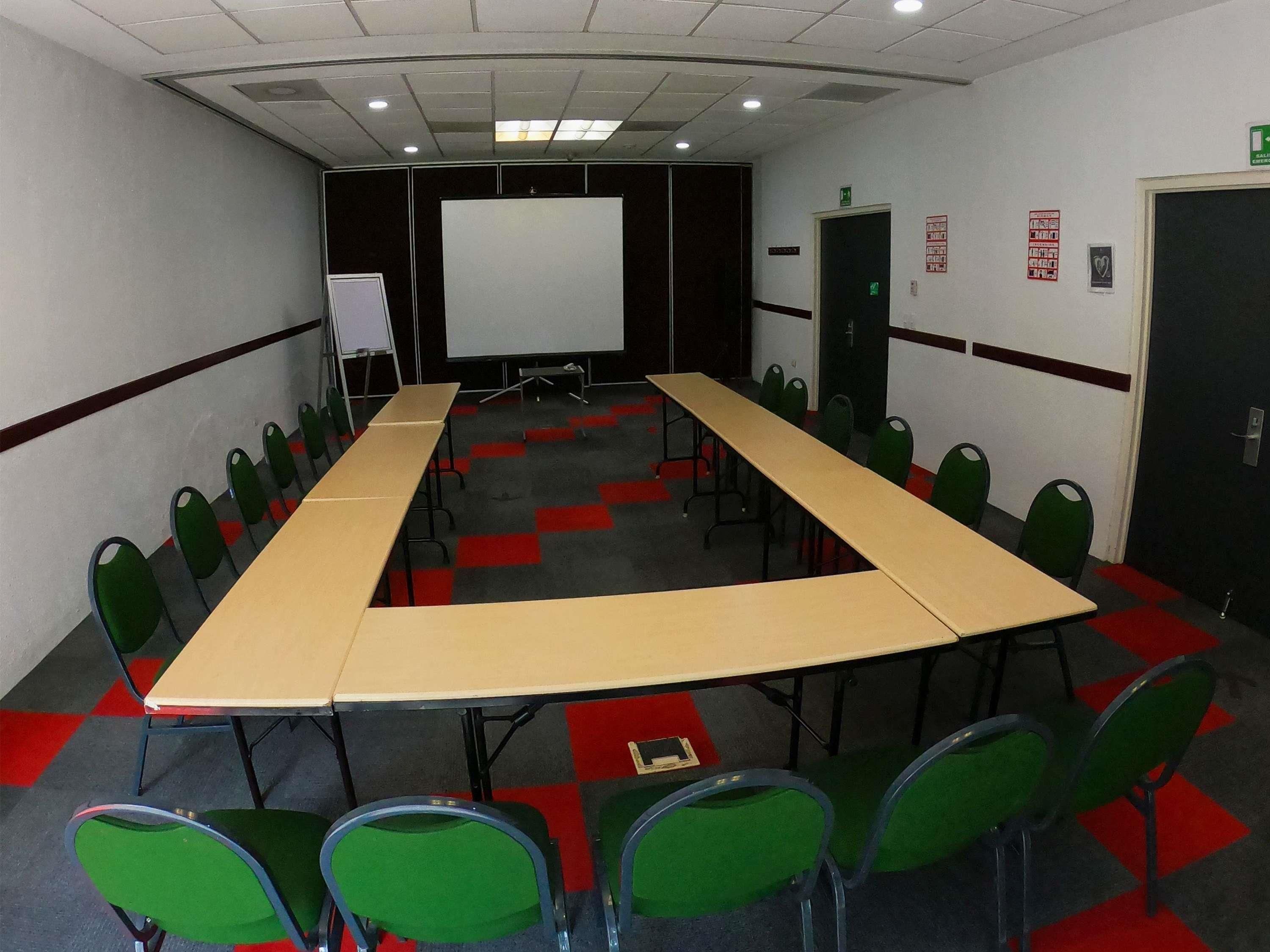 Una sala de conferencias con una larga mesa, sillas verdes, una pizarra y una pantalla de proyector.