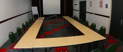 Una sala de conferencias con una larga mesa, sillas verdes, una pizarra y una pantalla de proyector.