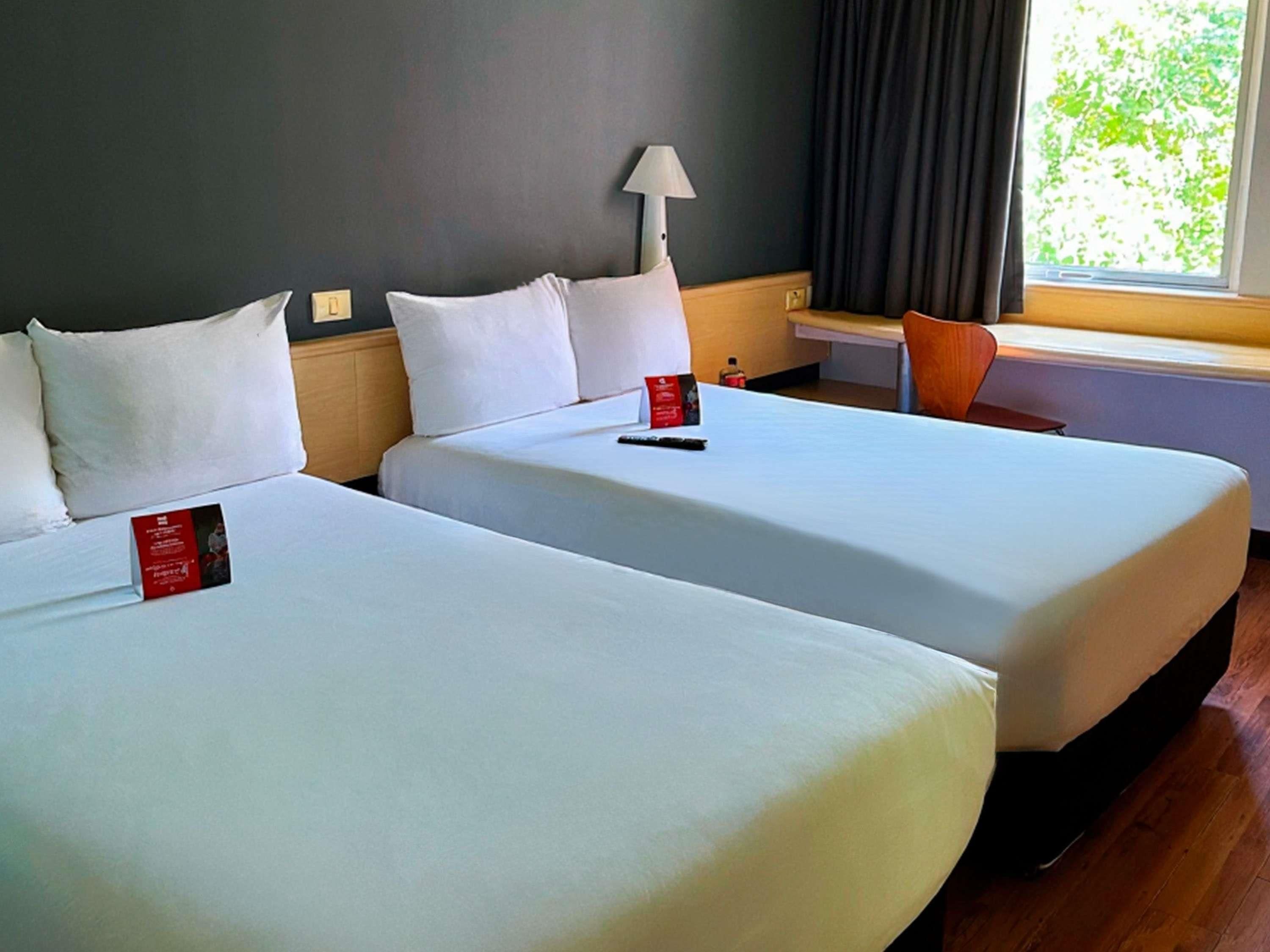 Two beds with white linens, a lamp, a window with curtains, and a wooden floor.