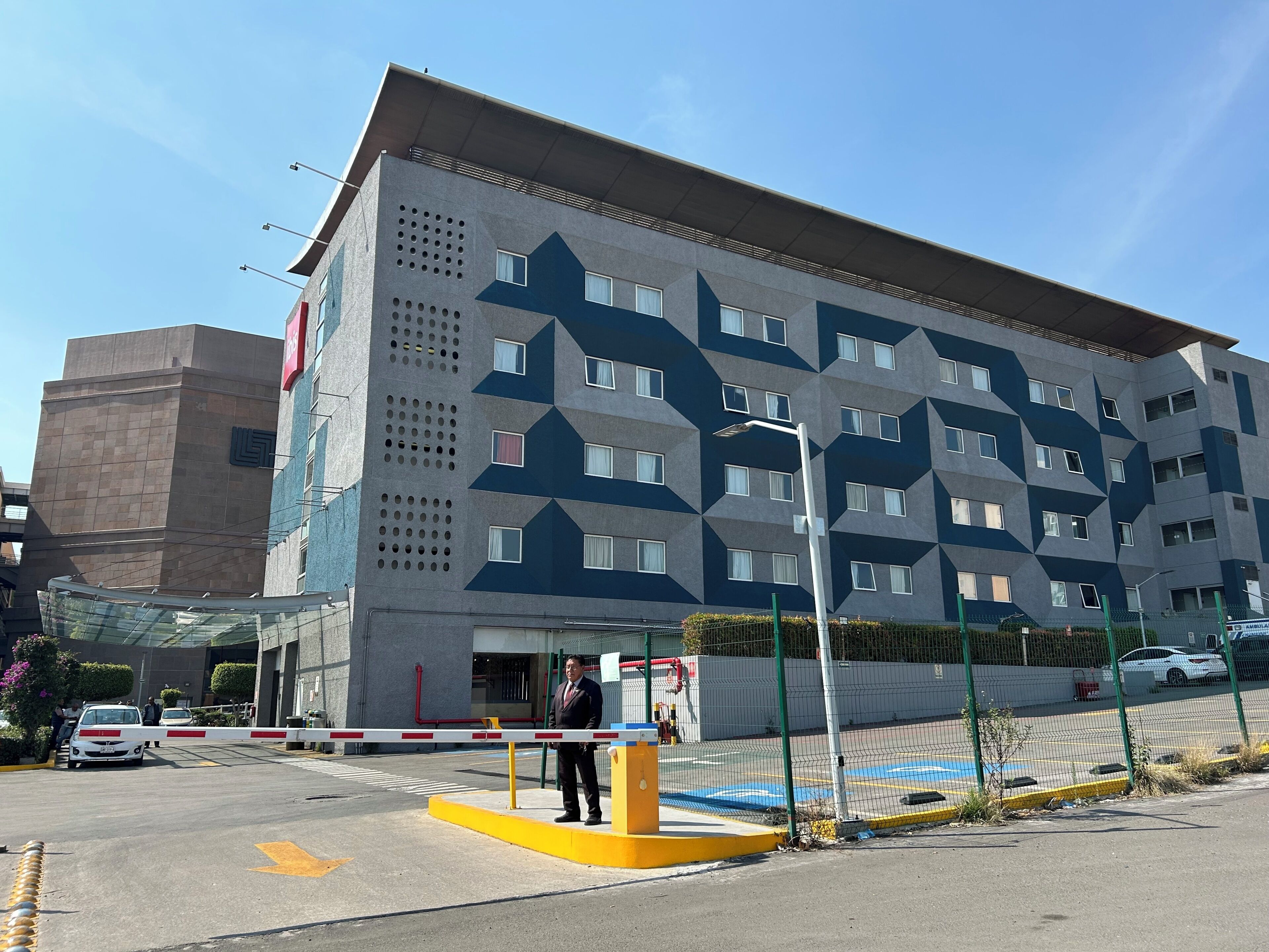 A multi-story hotel with a geometric facade, a gated entrance, and a parking area with marked spaces.